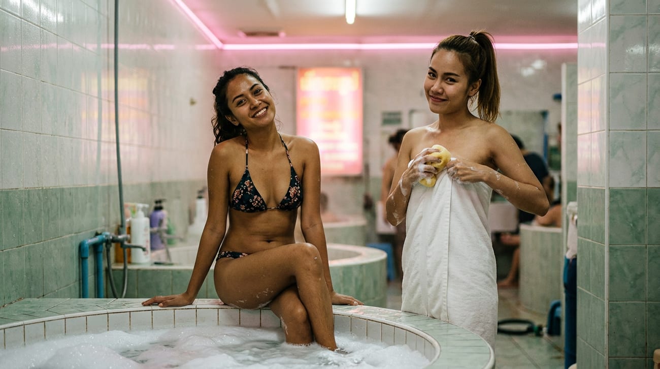 Two women at a tiled soapy massage bath, one in a floral bikini on the tub edge, one wrapped in a towel holding a sponge